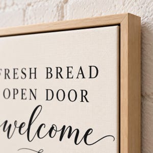 May include: A rectangular canvas wall art piece framed in light-colored wood. The off-white canvas features black text that reads FRESH BREAD, OPEN DOOR, and welcome in a mix of serif and script fonts, centered on the textured fabric surface.