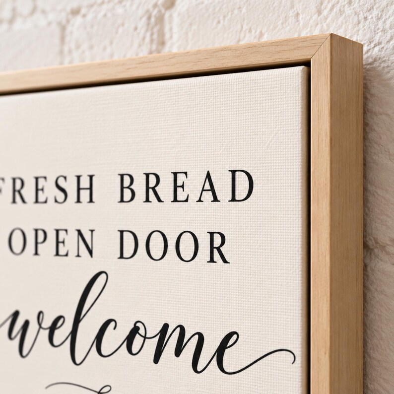 May include: A rectangular canvas wall art piece framed in light-colored wood. The off-white canvas features black text that reads FRESH BREAD, OPEN DOOR, and welcome in a mix of serif and script fonts, centered on the textured fabric surface.