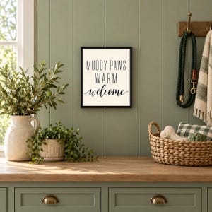 May include: A square wall sign with a black frame and a white background featuring the black text MUDDY PAWS WARM welcome. The sign is displayed on a sage green paneled wall above a wooden surface with plants and a woven basket.