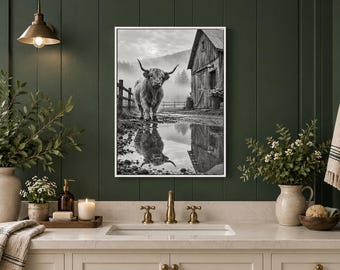 May include: A framed black and white wall art print featuring a Highland cow standing in a muddy field near a rustic wooden barn. The scene includes a puddle in the foreground reflecting the cow and the barn, set against a misty landscape.