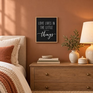 May include: A rectangular wall art piece in a light wood frame featuring a black background with the white text LOVE LIVES IN THE LITTLE things. The text is displayed in a combination of clean sans-serif capital letters and a cursive script.