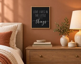 May include: A rectangular wall art piece in a light wood frame featuring a black background with the white text LOVE LIVES IN THE LITTLE things. The text is displayed in a combination of clean sans-serif capital letters and a cursive script.
