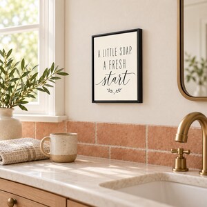 May include: A rectangular wall sign with a black frame and a cream-colored background. The text reads A LITTLE SOAP A FRESH start in black lettering, with a small decorative sprig graphic centered below the text.