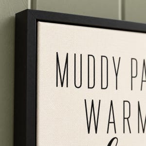 May include: A framed canvas wall art piece featuring a cream-colored background with black text. The visible portion of the text reads MUDDY PA and WARM. The canvas is enclosed in a thin, black, rectangular frame with a clean, sharp edge.