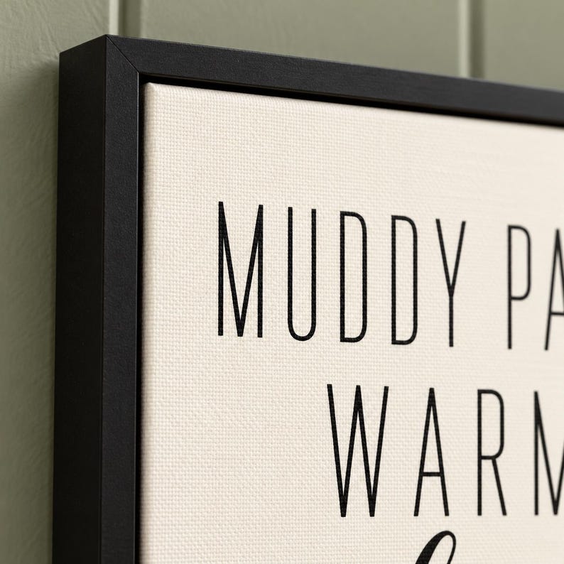 May include: A framed canvas wall art piece featuring a cream-colored background with black text. The visible portion of the text reads MUDDY PA and WARM. The canvas is enclosed in a thin, black, rectangular frame with a clean, sharp edge.