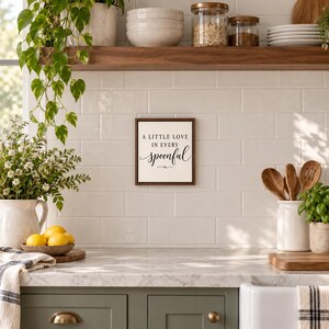 May include: A square wall sign with a dark wood frame and a white background featuring the black text A LITTLE LOVE IN EVERY spoonful, centered on a white tiled kitchen wall above a countertop with various kitchen items.