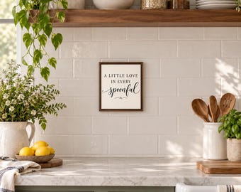 May include: A square wall sign with a dark wood frame and a white background featuring the black text A LITTLE LOVE IN EVERY spoonful, centered on a white tiled kitchen wall above a countertop with various kitchen items.
