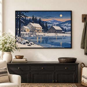 May include: A framed landscape painting depicting a snow-covered farmhouse and barn beside a frozen lake under a twilight sky with a full moon. The scene features dark evergreen trees, rolling hills in the distance, and warm light glowing from the house windows.