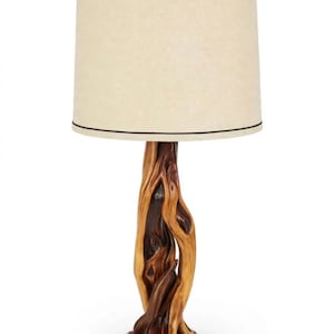 May include: A table lamp featuring a base crafted from a single piece of polished, gnarled natural wood with deep brown and tan grain patterns. The lamp is topped with a cylindrical, off-white fabric shade featuring thin black trim.