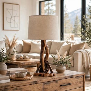 May include: A table lamp featuring a natural, twisted dark wood base with a circular wooden stand and a cylindrical beige fabric lampshade. The lamp is positioned on a wooden surface, showcasing the organic texture of the wood grain.