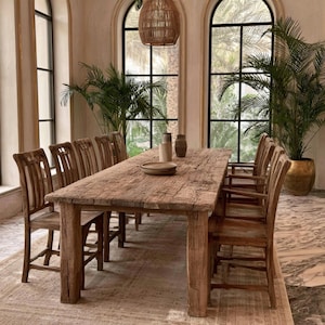 Puede incluir: Una mesa de comedor rectangular y larga hecha de madera de color marrón claro desgastada con una superficie rugosa y texturizada. La mesa está rodeada por ocho sillas de madera a juego con listones verticales en una habitación luminosa.