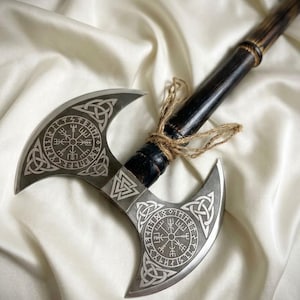 May include: A double-bitted axe with a dark wooden handle wrapped in twine. The metal head features intricate silver-toned engravings of circular Vegvisir symbols, triquetra knots, and a central Valknut emblem on both sides.