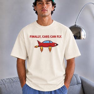 May include: A cream-colored short-sleeve t-shirt featuring a graphic of a red flying car with orange and yellow flames underneath. The text FINALLY, CARS CAN FLY. is printed in red above the car illustration. The shirt has a crew neckline.