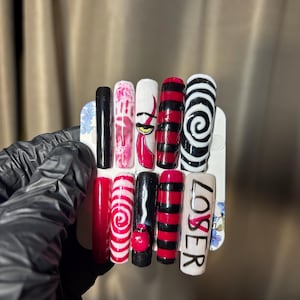 Puede incluir: Un juego de diez uñas postizas con varios diseños, incluyendo negro sólido, rayas rojas y negras, espirales rojas y blancas, una huella de mano roja, un ojo amarillo y la palabra LOSER escrita en letras negras sobre un fondo blanco.