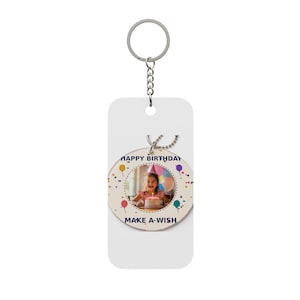 Puede incluir: Un llavero rectangular blanco con anilla y cadena de metal, que incluye un inserto acrílico circular. El inserto muestra una fotografía de un niño con un pastel de cumpleaños, rodeado de gráficos de globos de colores y el texto HAPPY BIRTHDAY MAKE A WISH.