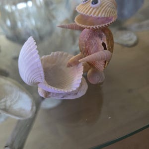 May include: A decorative figurine crafted from various seashells, featuring a creature-like form with googly eyes. The figure is positioned as if interacting with a separate, open, light-colored seashell that serves as a base or container.