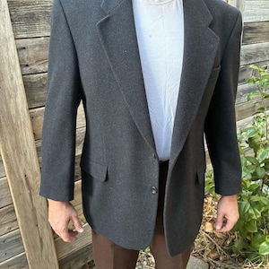 Könnte beinhalten: Ein anthrazitfarbener Blazer mit steigendem Revers, zwei Knöpfen auf der Vorderseite und zwei Pattentaschen. Der Blazer wird über einem schlichten weißen T-Shirt mit Rundhalsausschnitt getragen und mit einer braunen Hose kombiniert.