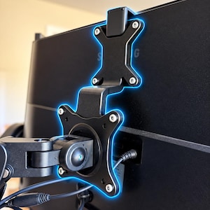 May include: A black metal monitor mount adapter bracket attached to the back of a black computer monitor. The adapter features a vertical cross-shaped design with four circular screw points and a central pivot arm connection point.
