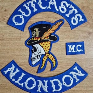 Könnte beinhalten: Ein Set aus gestickten Aufnähern mit einem blauen, bogenförmigen oberen Rocker mit dem Text OUTCASTS, einem blauen, bogenförmigen unteren Rocker mit dem Text N. LONDON, einem kleinen rechteckigen blauen Aufnäher mit M.C. und einem Totenkopf mit Feder.