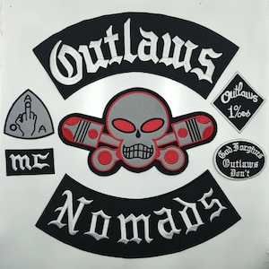 Op de afbeelding: Een verzameling geborduurde patches, waaronder een grote zwarte boog met witte tekst Outlaws, een centrale grijze schedel met rode ogen en gekruiste zuigers, een zwarte boog met witte tekst Nomads, en kleinere patches met tekst en symbolen.