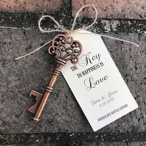May include: A copper-colored antique key bottle opener with a decorative design. The key has a tag attached with the text "The Key to Happiness is Love" and the names "Tara & Jesse" and the date "8.26.2017".