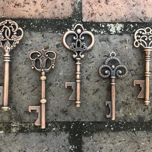 May include: Five antique-style bottle openers in the shape of keys. The keys are made of metal and have a bronze finish. Each key has a different design on the top, including a crown, a heart, and a flower.