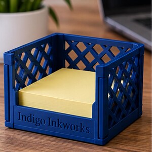 May include: A dark blue plastic desk organizer shaped like a miniature crate with a lattice pattern on the sides. The front panel features the embossed text Indigo Inkworks. A stack of square yellow sticky notes sits inside the container.