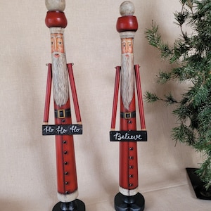 May include: Two tall, wooden Santa Claus figures. Each has a red coat, long white beard, and a small sign. One sign reads "Ho Ho Ho," and the other says "Believe." The figures have a round head and a black base.
