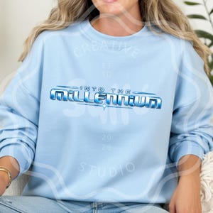 Op de afbeelding: Een lichtblauw sweatshirt met ronde hals en een grafische print op de borst met de tekst "INTO THE MILLENNIUM" in een metallic blauw, driedimensionaal lettertype met een steraccent op een effen lichtblauwe stof.