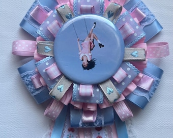 Puede incluir: Una escarapela circular con un botón central que muestra a una persona columpiándose boca abajo. La escarapela está hecha de cintas superpuestas en azul claro y rosa, algunas con lunares blancos, e incluye cintas colgantes.