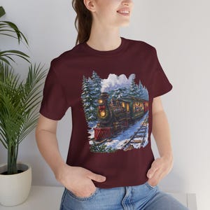 May include: A maroon short-sleeve t-shirt featuring a graphic print of a vintage steam locomotive traveling through a snowy forest. The train is decorated with a wreath and red bow, with smoke rising from the stack into a winter sky.
