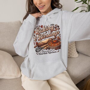 May include: A light grey hooded sweatshirt featuring a graphic print with the text Baking & Blessings, baked with love, and Heartfelt Kitchen. The design includes a rolling pin, a whisk, a pumpkin pie, and autumn leaves in warm tones.