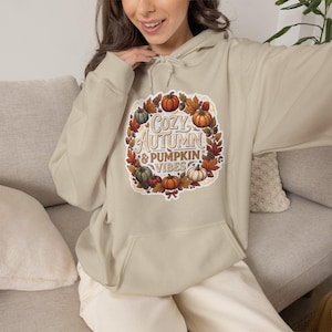 Puede incluir: Una sudadera con capucha de color beige que presenta un gráfico central de calabazas, hojas de otoño y bellotas dispuestas en una corona. El texto "COZY AUTUMN & PUMPKIN VIBES" está impreso en el centro con una fuente decorativa.