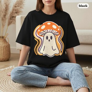 May include: A black short-sleeve t-shirt featuring a graphic of a white ghost wearing an orange mushroom cap with white spots. The design includes wavy yellow and purple lines surrounding the ghost. The word black is printed in the corner.