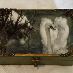 May include: A rectangular box with a hinged lid featuring a painted scene of white swans on dark water. The box has gold-toned metal filigree corner accents and a central metal latch. The sides of the box are a muted green color with gold trim.
