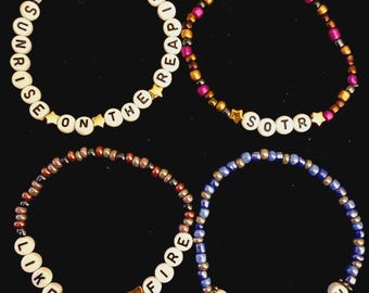 Puede incluir: Cuatro pulseras de cuentas con letras blancas y cuentas de vidrio de colores. Una pulsera dice SUNRISE ON THE REAPING, otra SOTR, otra LIKE ALL FIRE y otra HAYMITCH. Las pulseras incluyen pequeños detalles en forma de estrella dorada.