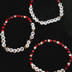 Puede incluir: Tres pulseras de cuentas con letras blancas que forman las frases ROCKY HORROR SHOW, DONT DREAM IT BE IT y TIME WARP. Las pulseras están hechas con pequeñas cuentas de semillas rojas, blancas y negras e incluyen separadores metálicos en forma de estrella.