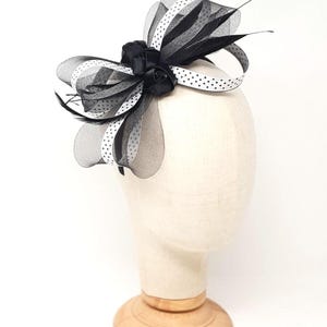 Può includere: Un copricapo bianco e nero con un design a fiocco. Il copricapo presenta rose nere, tessuto nero trasparente e nastro bianco a pois. Piume e sottili accenti neri aggiungono dettagli all'accessorio formale, esposto su una testa di manichino.