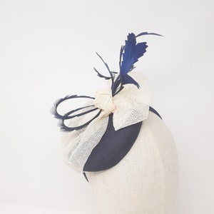May include: A navy blue and beige fascinator with a bow and feather accents. The headpiece is mounted on a headband. The bow is made of beige fabric and the feathers are navy blue.