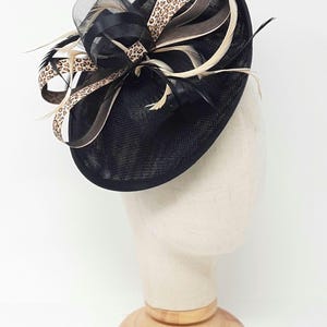 May include: A black hat with a mesh base, adorned with a bow featuring leopard print ribbon and feathers. The hat is displayed on a mannequin head, showcasing its design and structure. A stylish accessory.