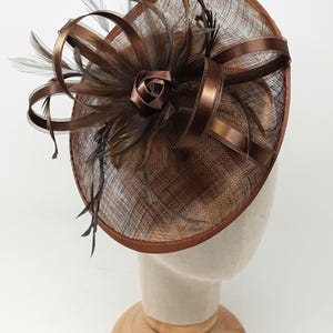 Könnte beinhalten: Ein brauner Fascinator-Hut mit kreisförmiger Basis, verziert mit einer Rose, Federn und Schleifen. Der Hut wird auf einem Mannequinkopf präsentiert. Die Farbpalette umfasst Braun- und Beigetöne und schafft ein elegantes Accessoire.