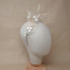 Könnte beinhalten: Ein cremefarbener Fascinator mit kreisförmiger Basis, verziert mit weißen Blumen, Perlen und Federn. Der Kopfschmuck ist an einem dünnen, silbernen Haarband befestigt. Das Design umfasst kleine, sternförmige Verzierungen.