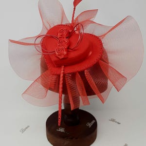 May include: A red hat with a heart-shaped crown, adorned with three rose accents and a sheer, flowing veil. The hat is displayed on a wooden stand, showcasing its intricate design and craftsmanship. The base of the stand is round and dark brown.