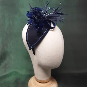 May include: A navy blue headpiece with a decorative arrangement of feathers, beads, and a velvet bow. The headpiece is mounted on a mannequin head, showcasing its design and structure. The base is a dark navy color with a lighter trim.