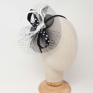 May include: A black fascinator headpiece featuring a circular base of black netting, a black ribbon with white polka dots, and white feathers. The accessory is mounted on a thin black headband, creating a structured and layered appearance.