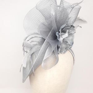 Può includere: Un fascinator grigio argento con un design a rete, un fiore di tessuto e piume. Il copricapo è montato su una sottile fascia per capelli. Il design presenta una rete e un tessuto a strati, creando un aspetto strutturato ed elegante.