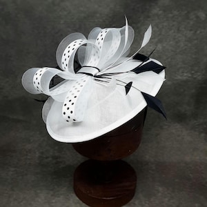 May include: A white fascinator hat featuring a circular base with a large bow made of white mesh ribbon with black polka dots. The design includes several thin, pointed black feathers extending outward from the center of the bow arrangement.