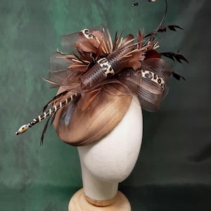 Op de afbeelding: Een bruine fascinator met een gladde, ronde basis versierd met bruine veren, donkerbruin gaas en een lint met luipaardprint. Een dunne, gebogen draad steekt omhoog, afgewerkt met kleine kralen en een lange, met kralen bezette stengel.
