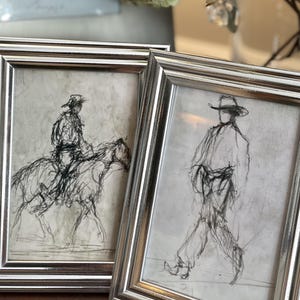 May include: Two rectangular silver-toned frames containing black ink sketches on white paper. The left frame displays a figure riding a horse, while the right frame shows a figure wearing a cowboy hat walking. Both artworks feature loose, expressive lines.