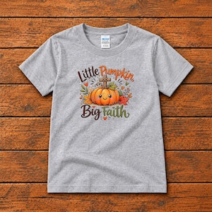 Puede incluir: Una camiseta gris jaspeado de manga corta con un gráfico de una calabaza naranja con cara sonriente y una cruz encima. El texto Little Pumpkin Big Faith está rodeado de hojas de otoño coloridas, bayas y pequeños corazones.